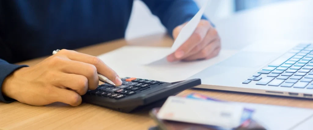 Finance professional reviewing invoices and calculating debt collection KPIs using a calculator and laptop.