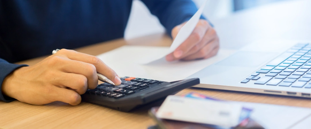 Finance professional reviewing invoices and calculating debt collection KPIs using a calculator and laptop.